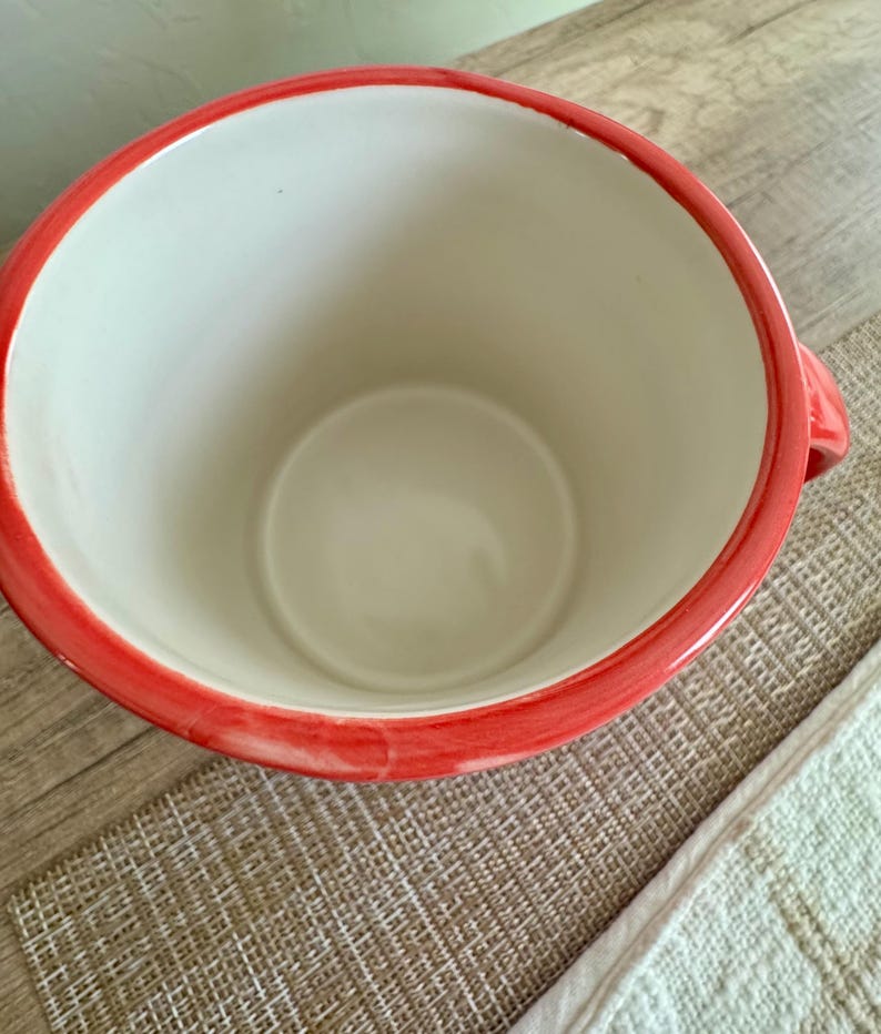 May include: A ceramic mug with a wide opening and a red rim. The interior is white, and the mug has a handle on the side. The mug is sitting on a textured surface.