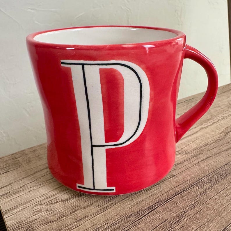 May include: Red ceramic mug with a white letter "P" in a bold, sans-serif font. The mug has a curved shape and a matching red handle. The interior of the mug is white. The mug is sitting on a wooden surface.