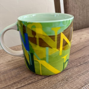 May include: A ceramic coffee mug with a green interior and handle. The exterior features a vibrant design with overlapping yellow, blue, and green letter shapes on a green background. The mug sits on a wooden surface.