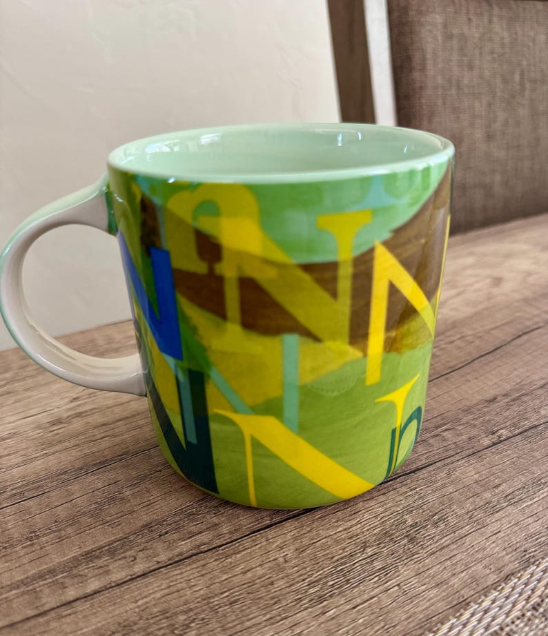 May include: A ceramic coffee mug with a green interior and handle. The exterior features a vibrant design with overlapping yellow, blue, and green letter shapes on a green background. The mug sits on a wooden surface.
