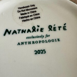 Puede incluir: Plato de cer&aacute;mica blanco con texto verde "Nathalie Lete" y "exclusivamente para ANTHROPOLOGIE". Una pegatina indica que el plato es solo para lavar a mano y est&aacute; hecho en China. El n&uacute;mero "2025" tambi&eacute;n est&aacute; impreso.
