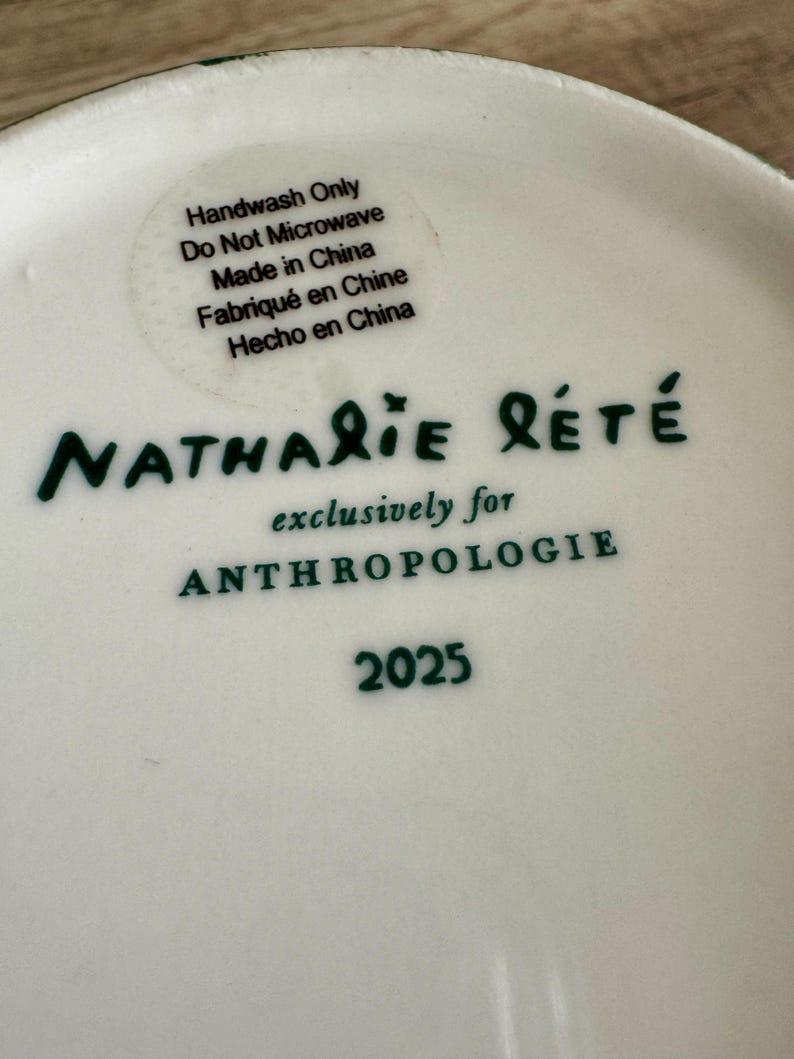 Puede incluir: Plato de cer&aacute;mica blanco con texto verde "Nathalie Lete" y "exclusivamente para ANTHROPOLOGIE". Una pegatina indica que el plato es solo para lavar a mano y est&aacute; hecho en China. El n&uacute;mero "2025" tambi&eacute;n est&aacute; impreso.