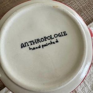 May include: Close-up of the base of a white ceramic mug with a red rim. The text "ANTHROPOLOGIE hand painted" is printed in black on the base of the mug. The mug is on a light-coloured surface.