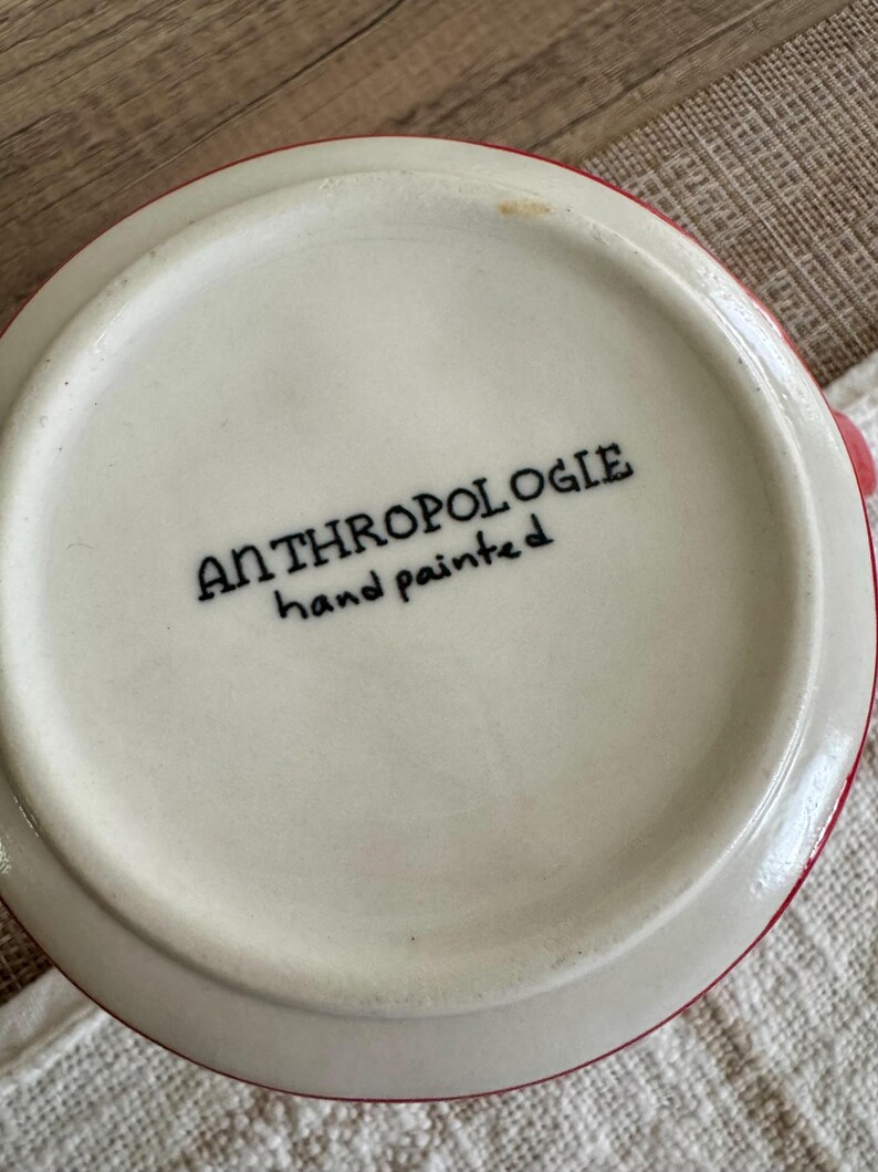 May include: Close-up of the base of a white ceramic mug with a red rim. The text "ANTHROPOLOGIE hand painted" is printed in black on the base of the mug. The mug is on a light-coloured surface.