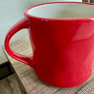 May include: A bright red ceramic mug with a white interior. The mug features a large handle and a glossy finish. The mug is on a wooden surface.