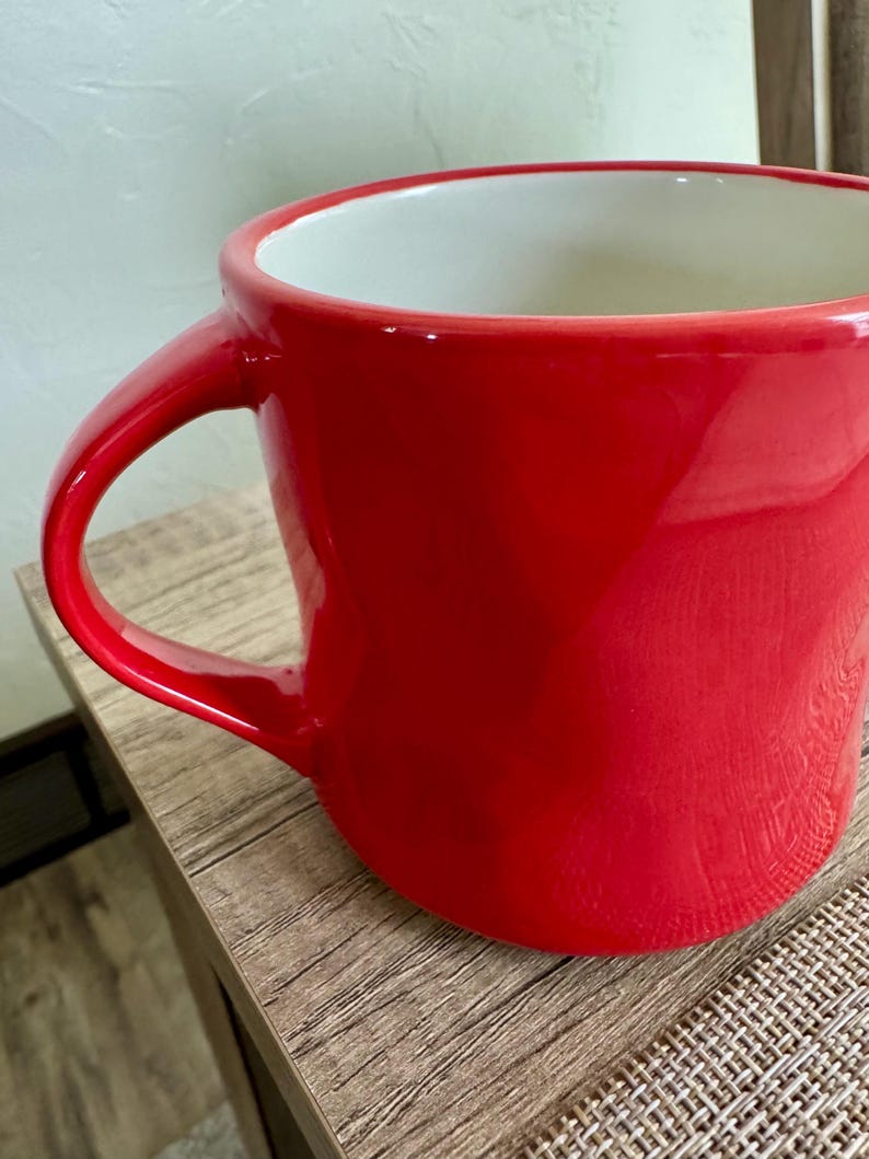 May include: A bright red ceramic mug with a white interior. The mug features a large handle and a glossy finish. The mug is on a wooden surface.