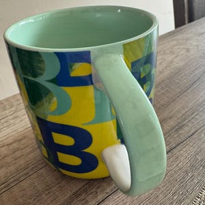 May include: A green ceramic mug with a yellow, blue, and green abstract design. The mug has a large handle and is decorated with the letters "B" and "E".