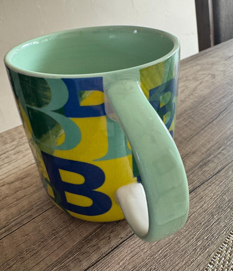 May include: A green ceramic mug with a yellow, blue, and green abstract design. The mug has a large handle and is decorated with the letters "B" and "E".