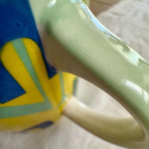 May include: Close-up of a ceramic mug with a light green handle and a body decorated with blue, yellow, and light green geometric shapes. The mug has a glossy finish and a white interior.