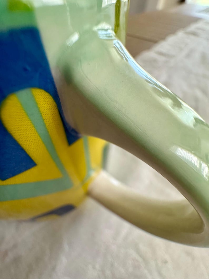 May include: Close-up of a ceramic mug with a light green handle and a body decorated with blue, yellow, and light green geometric shapes. The mug has a glossy finish and a white interior.
