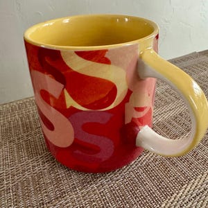 May include: A ceramic mug with a yellow interior and handle. The exterior is red with overlapping letter "S" designs in shades of yellow and pink. The mug sits on a woven surface.