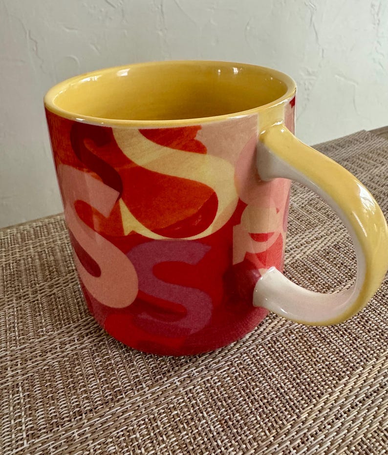 May include: A ceramic mug with a yellow interior and handle. The exterior is red with overlapping letter "S" designs in shades of yellow and pink. The mug sits on a woven surface.