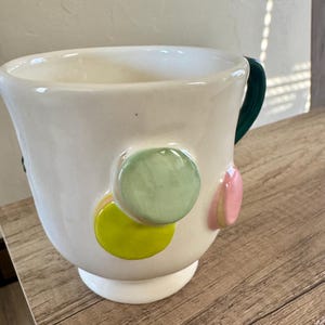 K&ouml;nnte beinhalten: Eine wei&szlig;e Keramiktasse mit gr&uuml;nem Henkel und bunten, runden, erhabenen Verzierungen. Die Verzierungen umfassen gr&uuml;ne, gelbe und rosa Kreise. Die Tasse steht auf einer Holzoberfl&auml;che.