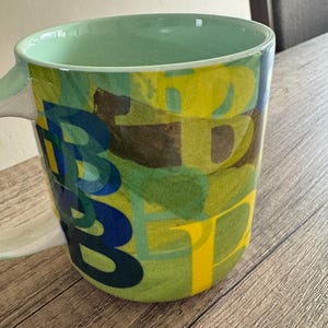 May include: A green ceramic mug with a colorful abstract design featuring blue, yellow, and brown letters. The mug has a white handle.