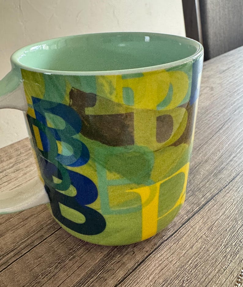May include: A green ceramic mug with a colorful abstract design featuring blue, yellow, and brown letters. The mug has a white handle.