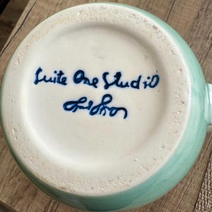 Pode incluir: Uma caneca de cer&acirc;mica com exterior verde-&aacute;gua claro e interior branco. A base da caneca apresenta texto azul manuscrito que diz "Suite One Studio Lisbon". A caneca est&aacute; sobre uma superf&iacute;cie de madeira.