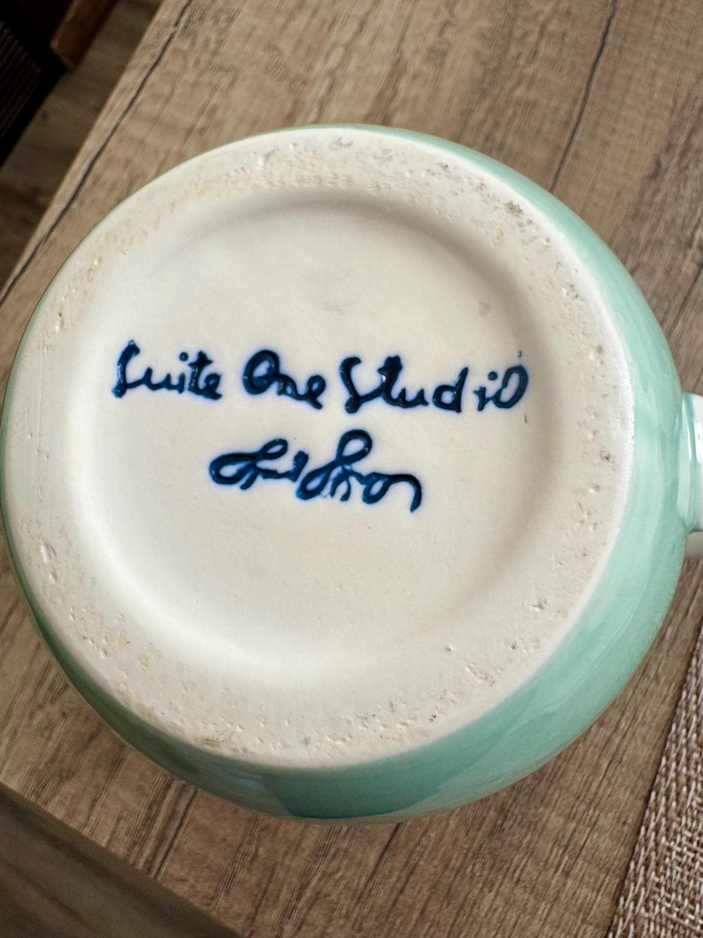 Pode incluir: Uma caneca de cer&acirc;mica com exterior verde-&aacute;gua claro e interior branco. A base da caneca apresenta texto azul manuscrito que diz "Suite One Studio Lisbon". A caneca est&aacute; sobre uma superf&iacute;cie de madeira.