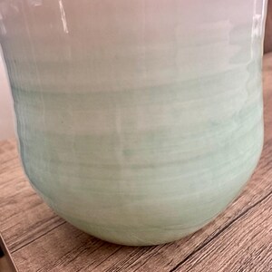 Pode incluir: Um vaso de cer&acirc;mica grande com um acabamento liso e brilhante. O vaso faz a transi&ccedil;&atilde;o de um branco suave no topo para um verde suave na base, com um efeito sutil de pincelada horizontal. O vaso tem uma forma arredondada e bulbosa.