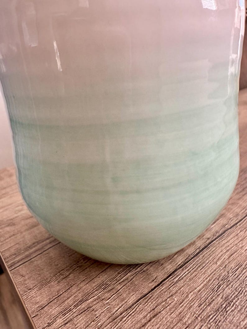 Pode incluir: Um vaso de cer&acirc;mica grande com um acabamento liso e brilhante. O vaso faz a transi&ccedil;&atilde;o de um branco suave no topo para um verde suave na base, com um efeito sutil de pincelada horizontal. O vaso tem uma forma arredondada e bulbosa.