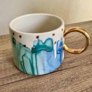 May include: A white ceramic mug featuring a watercolor-style abstract pattern in shades of blue and teal. The exterior is decorated with small, gold-colored star-shaped accents. The mug includes a round, metallic gold-finished handle on the side.
