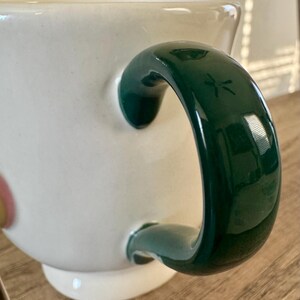 K&ouml;nnte beinhalten: Eine wei&szlig;e Keramiktasse mit einem gl&auml;nzenden, dunkelgr&uuml;nen Henkel. Der Henkel hat ein eingraviertes Libellen-Design. Die Tasse steht auf einer Holzoberfl&auml;che.