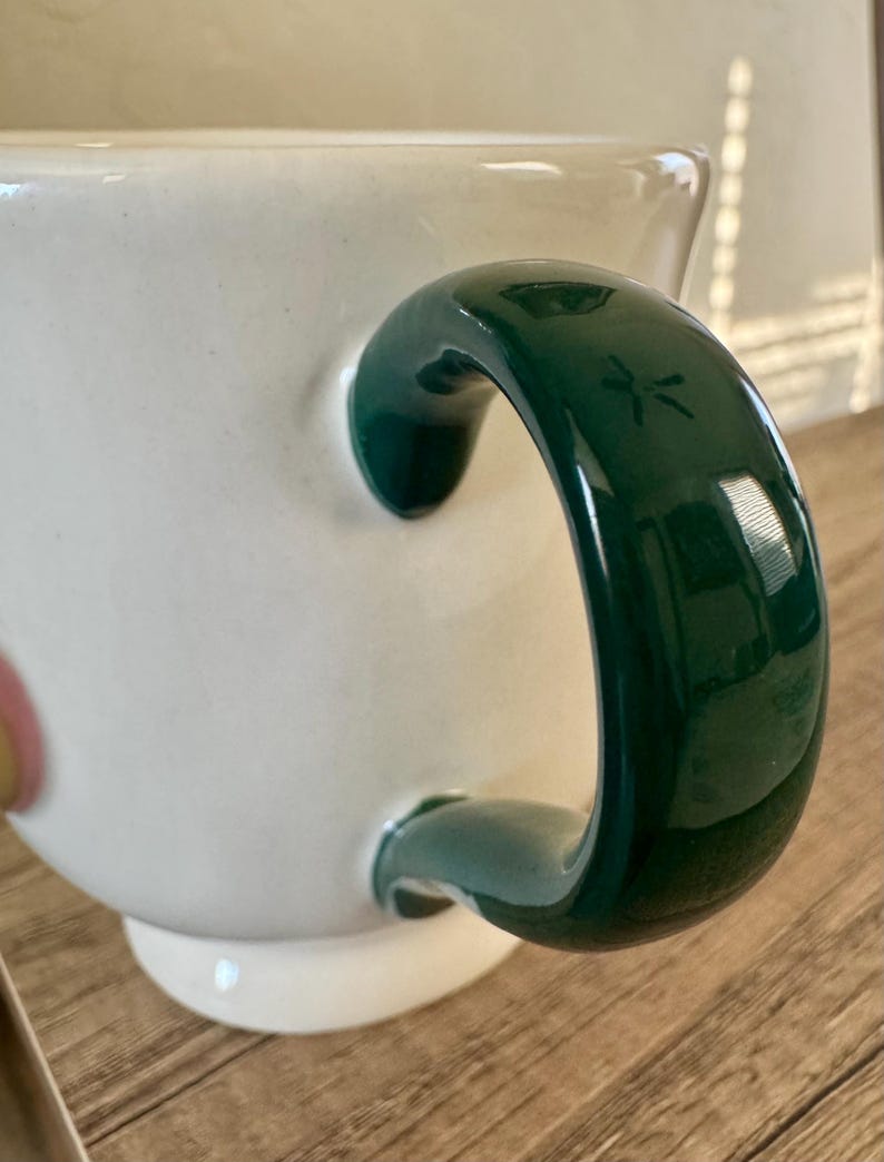 K&ouml;nnte beinhalten: Eine wei&szlig;e Keramiktasse mit einem gl&auml;nzenden, dunkelgr&uuml;nen Henkel. Der Henkel hat ein eingraviertes Libellen-Design. Die Tasse steht auf einer Holzoberfl&auml;che.