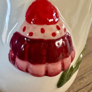 K&ouml;nnte beinhalten: Nahaufnahme einer wei&szlig;en Keramiktasse mit einem erhabenen, farbenfrohen Cupcake-Design. Der Cupcake hat eine rote Kirsche oben, eine rosa und rote Zuckerguss-Schicht und eine wei&szlig;e Basis. Gr&uuml;ne Blattakzente sind an der Seite der Tasse zu sehen.