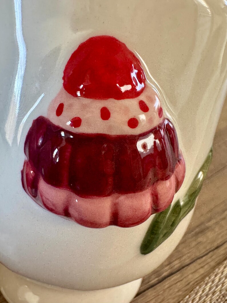K&ouml;nnte beinhalten: Nahaufnahme einer wei&szlig;en Keramiktasse mit einem erhabenen, farbenfrohen Cupcake-Design. Der Cupcake hat eine rote Kirsche oben, eine rosa und rote Zuckerguss-Schicht und eine wei&szlig;e Basis. Gr&uuml;ne Blattakzente sind an der Seite der Tasse zu sehen.