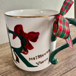 Puede incluir: Taza de cer&aacute;mica blanca con borde dorado, decorada con un lazo rojo y detalles verdes. La taza tiene un asa verde y una cinta a cuadros roja, verde y blanca atada. Las palabras "Meilleurs Voeux" est&aacute;n escritas en la taza.