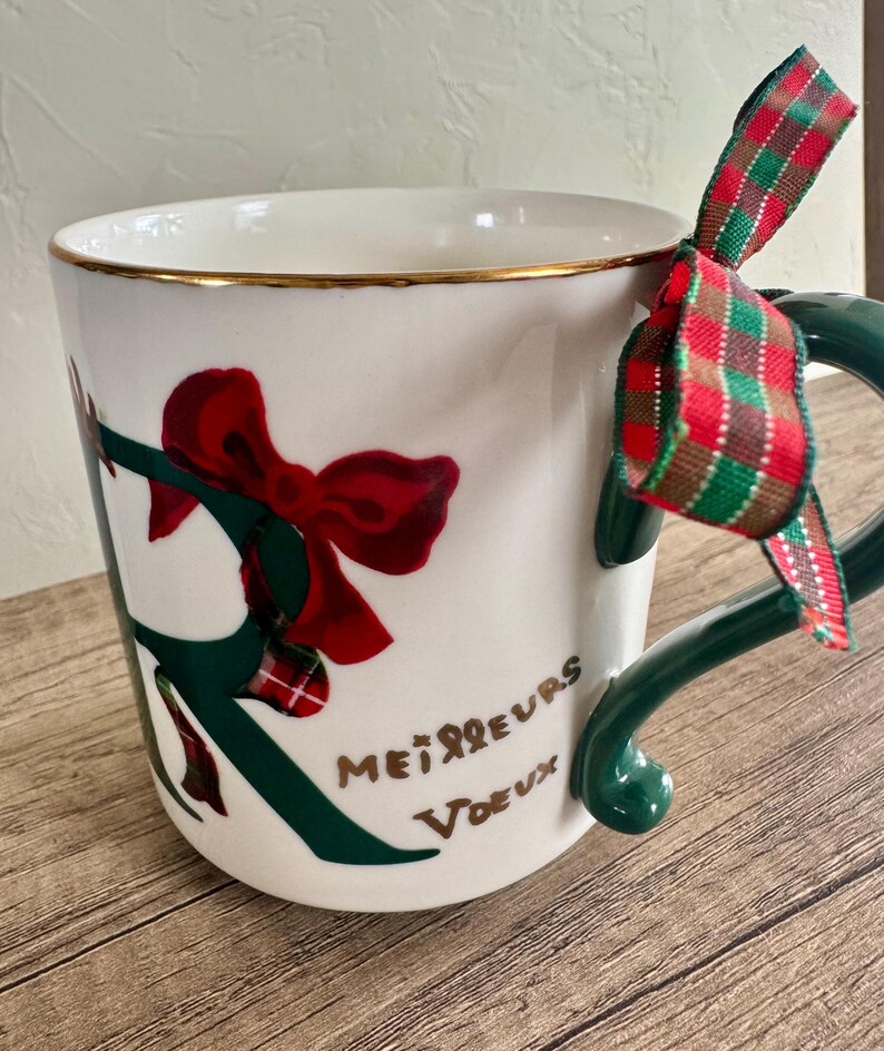Puede incluir: Taza de cer&aacute;mica blanca con borde dorado, decorada con un lazo rojo y detalles verdes. La taza tiene un asa verde y una cinta a cuadros roja, verde y blanca atada. Las palabras "Meilleurs Voeux" est&aacute;n escritas en la taza.
