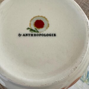 May include: Cream ceramic bowl with a floral pattern on the rim. The base of the bowl has a red and gold circular design and the text "by ANTHROPOLOGIE" in black.