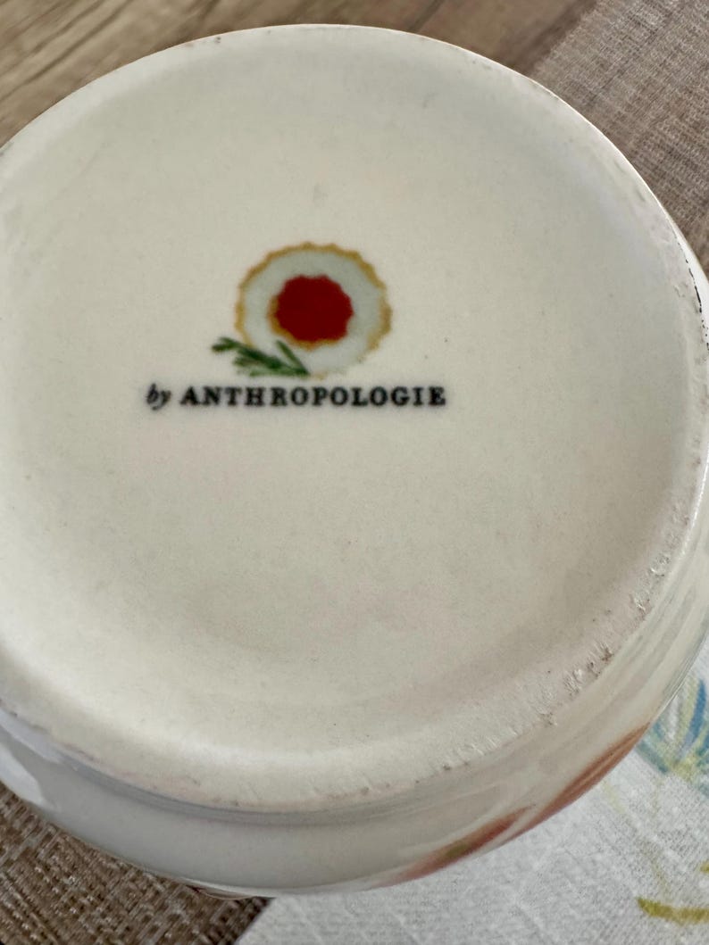 May include: Cream ceramic bowl with a floral pattern on the rim. The base of the bowl has a red and gold circular design and the text "by ANTHROPOLOGIE" in black.