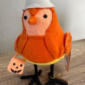 May include: A plush orange bird figurine with a white hat and black legs. The bird is holding a small orange trick-or-treat bag with a jack-o'-lantern face. A yellow band encircles the bird's midsection. A Halloween decoration.