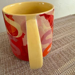 May include: A ceramic mug with a yellow interior and handle. The exterior features a colorful abstract design in shades of red, orange, and pink. The mug sits on a textured, neutral-toned surface.