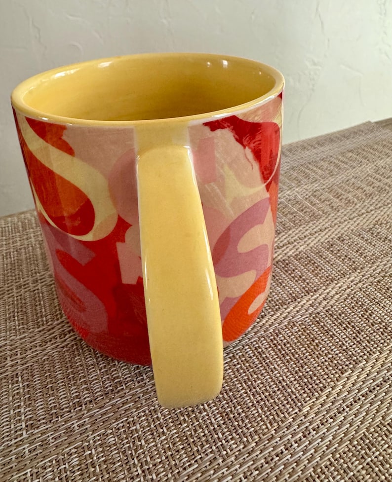 May include: A ceramic mug with a yellow interior and handle. The exterior features a colorful abstract design in shades of red, orange, and pink. The mug sits on a textured, neutral-toned surface.
