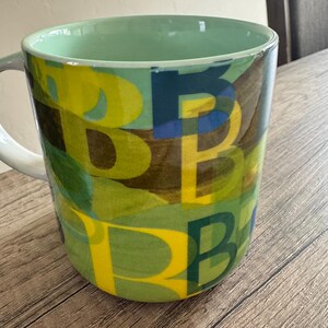 May include: A green ceramic mug with a yellow, blue, and green abstract pattern. The pattern is made up of overlapping letters.