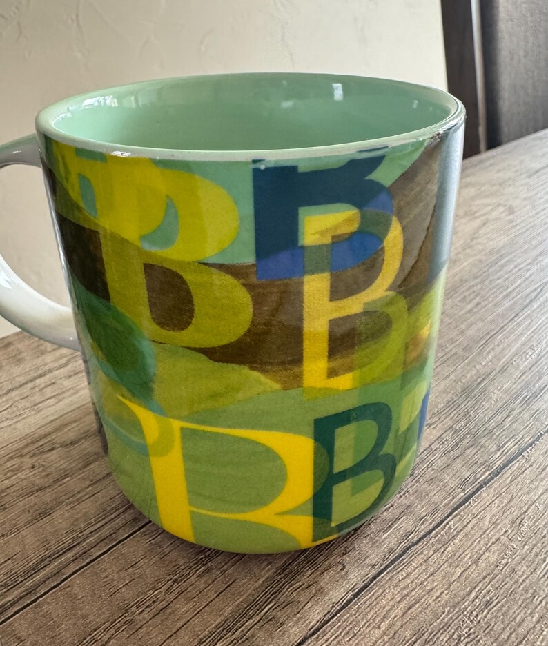 May include: A green ceramic mug with a yellow, blue, and green abstract pattern. The pattern is made up of overlapping letters.
