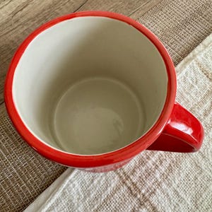 May include: A red ceramic mug with a white interior. The mug has a red rim and a matching red handle. The mug is on a white and tan textured surface.