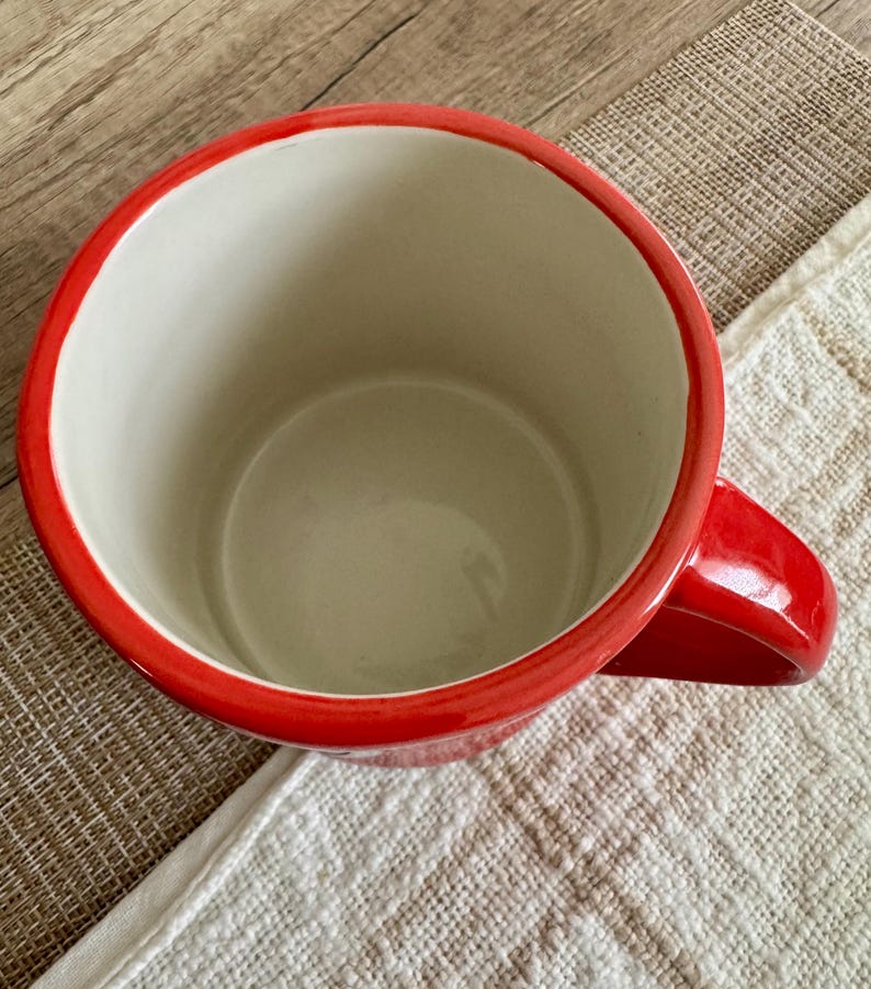 May include: A red ceramic mug with a white interior. The mug has a red rim and a matching red handle. The mug is on a white and tan textured surface.
