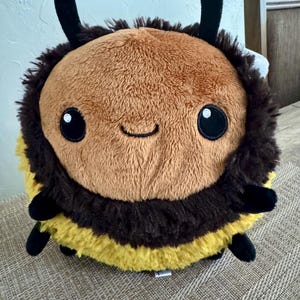 May include: A plush bumblebee toy with a round body. The toy features a brown head, black and yellow striped body, black antennae, and small black legs. The bee has large, round black eyes and a smiling mouth.