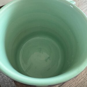 May include: A light green ceramic mug with a white interior. The mug is empty and sitting on a wooden surface.