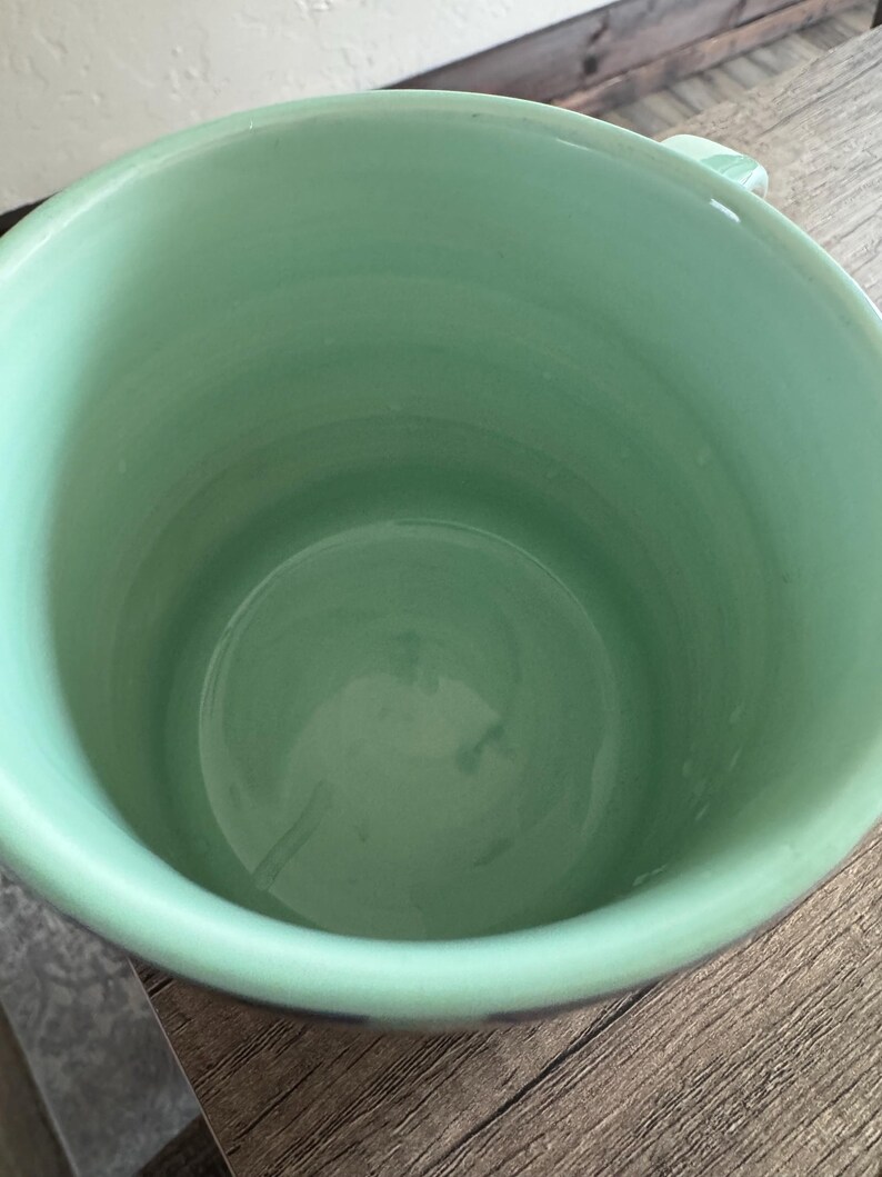 May include: A light green ceramic mug with a white interior. The mug is empty and sitting on a wooden surface.