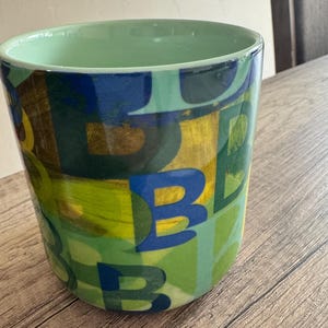 May include: A green ceramic mug with a colorful abstract design featuring the letter "B" repeated in various colors and sizes.