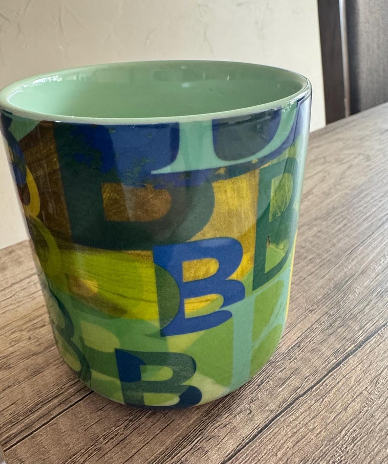 May include: A green ceramic mug with a colorful abstract design featuring the letter "B" repeated in various colors and sizes.