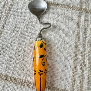 May include: A yellow ceramic spoon with a black and green floral design. The spoon has a metal handle that is bent into a hook shape.