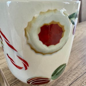 May include: Cream ceramic mug decorated with festive holiday designs. The mug features a large, round cookie with a red centre, candy canes, and other colourful holiday treats. Ideal for enjoying hot beverages during the festive season.