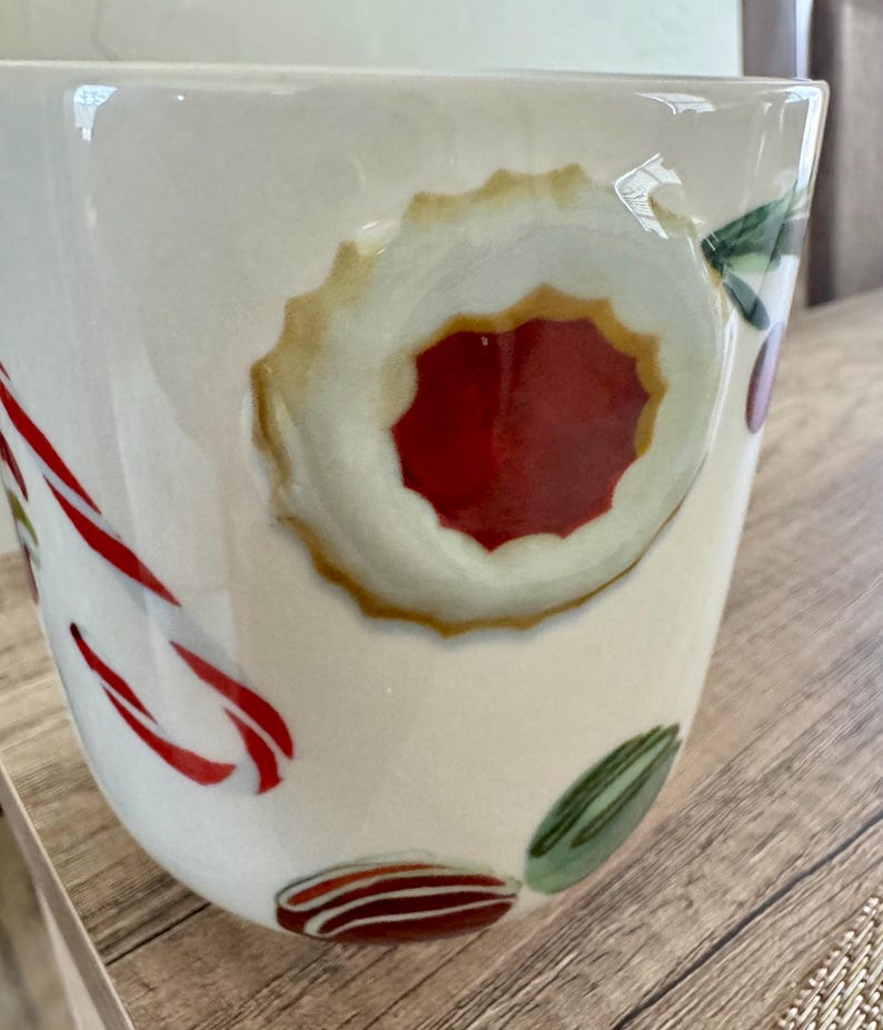 May include: Cream ceramic mug decorated with festive holiday designs. The mug features a large, round cookie with a red centre, candy canes, and other colourful holiday treats. Ideal for enjoying hot beverages during the festive season.