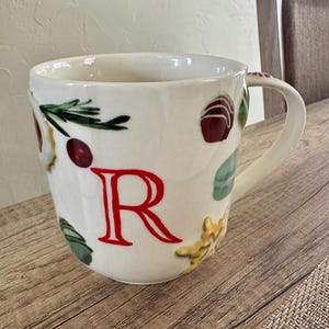 May include: White ceramic mug with a large red letter "R" and decorative food illustrations. The mug features a handle and is adorned with images of berries, herbs, and pastries. The mug sits on a wooden surface.