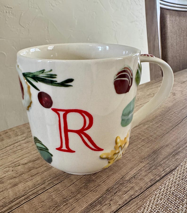 May include: White ceramic mug with a large red letter "R" and decorative food illustrations. The mug features a handle and is adorned with images of berries, herbs, and pastries. The mug sits on a wooden surface.