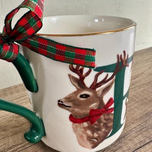 Puede incluir: Taza de cer&aacute;mica blanca con asa verde y borde dorado. La taza presenta una ilustraci&oacute;n de ciervo con un lazo rojo y la letra "E". Una cinta a cuadros roja, verde y dorada est&aacute; atada alrededor del asa.